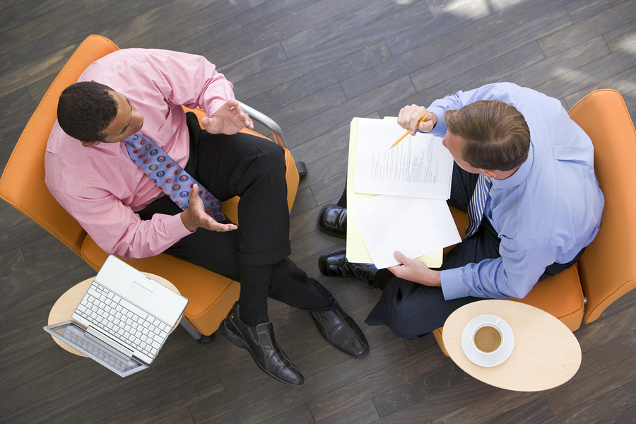Two men in armchairs discussing paperwork with a laptop nearby